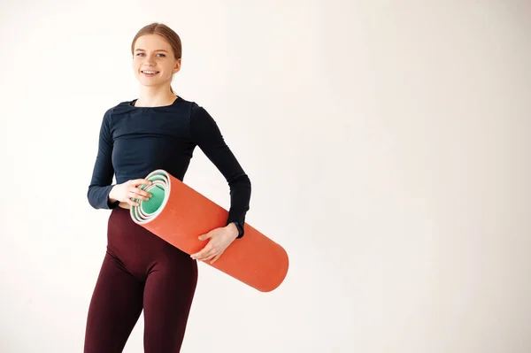 年轻强壮的运动健美教练女教练穿着深色运动服 花时间呆在家里的体育馆里 站在素净的白色背景上 拿着瑜伽垫 — 图库照片