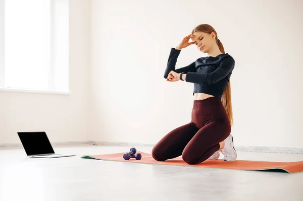 运动后在瑜伽垫上休息的疲惫女人 疲惫疲惫的女运动员带着两个哑铃坐在体育馆的地板上 测量健身手镯的压力 — 图库照片