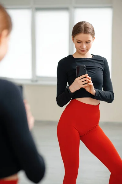 赤いタイトなレギンスとブラックトップの魅力的な女性は パーソナルスマートフォン上のミラーで自撮りを作ります 明るいジムでスリムなボディ形状のトレーニングを持つ若い女性 近代技術とスポーツの概念 — ストック写真
