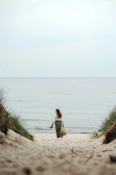一个有着棕色长发的女士带着鞋子和编织的袋子去沙滩的背景图 穿便衣的年轻女子独自在海滨度假 — 图库照片