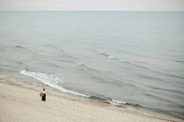 壮丽的大海和沿着荒芜的海岸漫步的女士的头像 穿着长裙和白衬衫的女人手提包走在沙滩上 享受着宁静和大海的喧嚣 — 图库照片