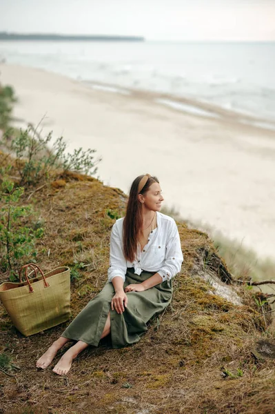 漂亮的年轻女性穿着橄榄绿长裙和白衬衫坐在地上观看海浪 拥有一头棕色长发和米色头巾的快乐女士 双手放在膝上 温和地微笑着 — 图库照片