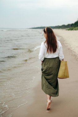Elinde büyük bir plaj çantasıyla kumlu sahilde gezinen koyu saçlı bir kadının arka görüntüsü. Sıradan giyinmiş bir kadın dalgaları seyrediyor ve boş bir adada yürümekten zevk alıyor. Uyum ve ilham kavramı.