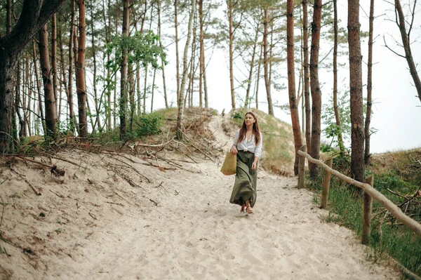 美丽的年轻女子走在沙滩小径上 提着大麻袋 她的眼前尽收眼底 身穿深绿色长裙和白衬衫的瘦瘦的女士欣赏森林风光 — 图库照片