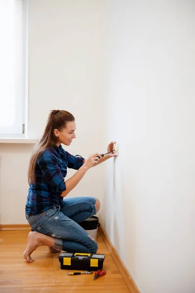 Ekose gömlekli genç esmer kadın tornavidayla elektrik prizini tamir ediyor. Duvara yeni soket takılıyor