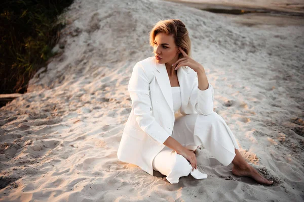 年轻女子金发碧眼 坐在沙滩上 在关于自然的专业摄影课上俯视一旁 穿着白色西服的女模特站在室外相机前 — 图库照片