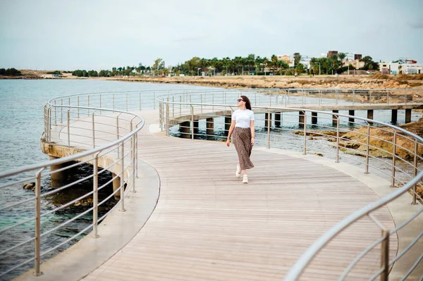 迷人的年轻女子戴着太阳镜 穿着夏装走在海滨附近的木制码头上 沙滩和水面上的波浪 — 图库照片