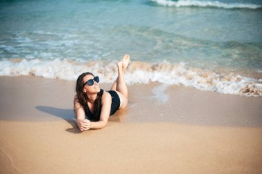 Modaya uygun güneş gözlüklü ve turkuaz renkli suyun yanındaki kumlu sahilde mayo giyen mutlu ve çekici bir kadın. Yaz zamanı, tatil ve insanlar konsepti.