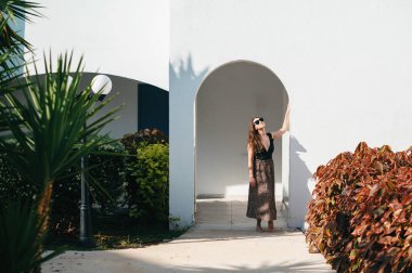 Hoş, koyu renk saçlı, güneş gözlüklü ve şık elbiseli bir kadın otel odalarına giriyor. Yaz tatili için lüks tatil beldesi.