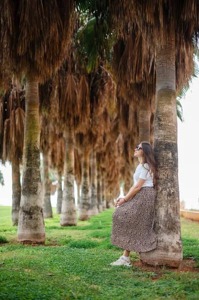站在棕榈树中间时 戴着太阳镜 穿着时髦衣服的漂亮年轻女子面带微笑地看着相机 在异国情调的度假胜地享受暑假的女游客 — 图库照片