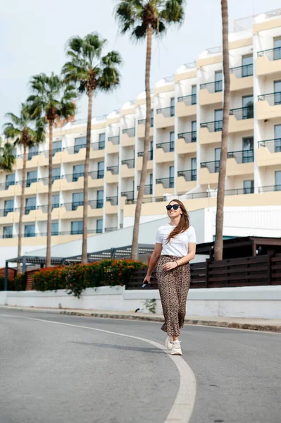 穿着太阳镜和时髦衣服的年轻女子带着棕榈树漫步在现代酒店的地盘上 热带度假胜地暑假 — 图库照片