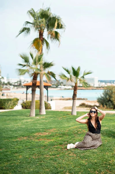 迷人的高加索女人 戴着太阳镜 穿着夏装 坐在棕榈树附近的草地上 享受奢华的异国情调 享受和放松的概念 — 图库照片
