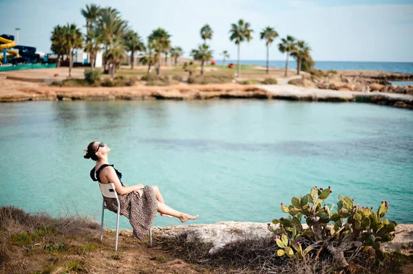 戴着太阳镜 身穿夏装的美丽的高加索女人坐在海滨的椅子上 手心紧握 年轻快乐的黑发姑娘在异国情调的度假胜地度假 — 图库照片