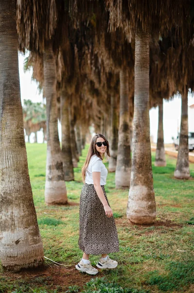 站在棕榈树中间时 戴着太阳镜 穿着时髦衣服的漂亮年轻女子面带微笑地看着相机 在异国情调的度假胜地享受暑假的女游客 — 图库照片