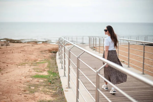 迷人的年轻女子戴着太阳镜 穿着夏装走在海滨附近的木制码头上 沙滩和水面上的波浪 — 图库照片