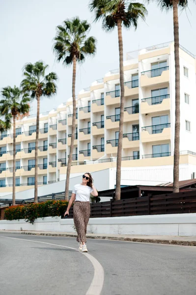 穿着太阳镜和时髦衣服的年轻女子带着棕榈树漫步在现代酒店的地盘上 热带度假胜地暑假 — 图库照片