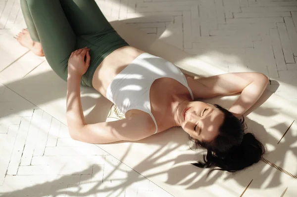 白种人黑发女士躺在室内阳光下的白色健身垫上 穿着舒适运动服的无忧无虑的女性 闭上眼睛练习瑜伽 放松身心 — 图库照片
