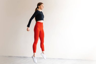 Aktif sağlıklı bir kadın kardiyo egzersizleri yapıyor ve beyaz stüdyonun pencerelerinden atlıyor. Dar spor kıyafetleri giymiş, bacak kaslarını çalıştıran enerjik bir kadın..