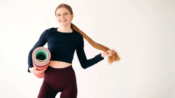 照片上 一个快乐的健身女士拿着橡胶垫子站在镜头前 在白色背景上开心地笑着 成功锻炼后 情绪激动的女性手牵着马尾辫 感到满意 — 图库照片