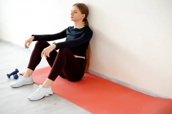 在健身中心的地板上用运动器材进行了紧张的锻炼后 筋疲力尽的女性正在休息 年轻的女运动员坐在红色的健身垫上 闭上了眼睛 恢复了体力 — 图库照片