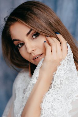 Beyaz lüks gelinlik içinde tatlı ve şehvetli esmer gelin. Mavi kumaş arka planlı çatı katındaki kameraya bakıyor. 