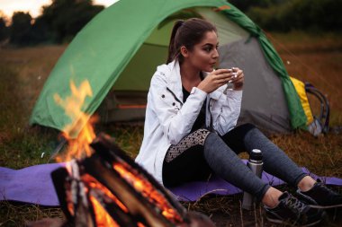 Smiling young woman drinking hot tea near the bonfire outside. Woman drinking a cup of warm tea alone near the tent