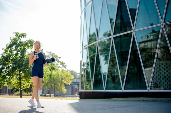 トレーニング後にヨガマットを携帯し 路上に立ちながら携帯電話を使用するスリムスポーツ女性 スポーツコンセプト — ストック写真