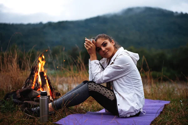 たき火の近くに座っている間 白人女性観光客が熱いお茶を飲むの側面図 キャンプ中に川や緑の自然を眺めながら素敵な女性 — ストック写真