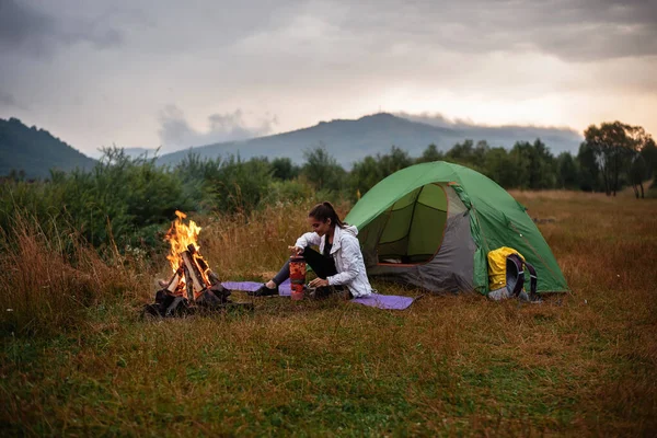 Full length view of the smiling young woman preparing putting the kettle on the fire. Joyful woman preparing drink outside. Woman drinking a cup of warm tea 