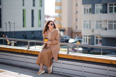 Açık hava moda detayları. Genç esmer kadın şehirde bir bankta otururken bir fincan kahvenin tadını çıkarıyor. Güneşli bahar günü.