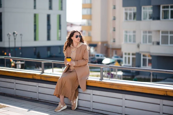 アウトドアファッションの詳細 コートを着た若いブルネットの女性は 街のベンチに座っている間にコーヒーを楽しんでいます 春の晴れた日 — ストック写真