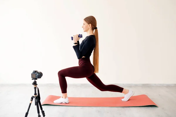Professional personal trainer recording video on camera during physical activity for own followers. Flexible fitness woman doing leg workout exercises and practicing lunges with dumbbells on mat.