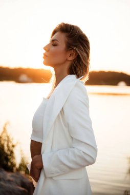 Göl kenarındaki profesyonel fotoğraf çekimi sırasında kameraya kapalı bakışlı şehvetli genç bir kadının yan görüntüsü. Sarışın, tasarımcı beyaz takım elbiseli bir kadın model..