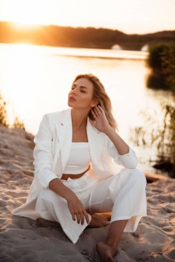 Göl kenarında kumların üzerinde oturan şık beyaz takım elbiseli etkili genç sarışın. İnanılmaz günbatımının bulanık arkaplanı. Moda ve güzellik kavramı.