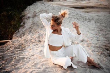 Güzel, beyaz takım elbiseli bir bayan kumların üzerinde oturuyor, sarı saçlara dokunuyor ve yan bakıyor. Mutlu kadın gün batımında doğada fotoğraf çekimi yapıyor..