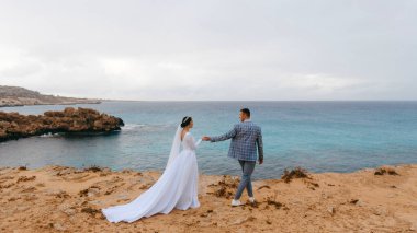 Mavi deniz kıyısındaki kayalık bir sahilde yürüyen genç bir düğün çifti. Damat gelinin elini tutuyor. Güzel düğün günü.