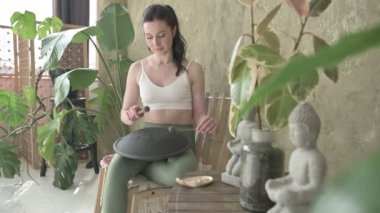Yeşil dar tayt ve beyaz bluz giymiş güzel bir bayan evde tank davuluyla pratik yapıyor. Koyu saçlı, at kuyruklu, dizlerinde müzik aleti olan ve ses çıkarmaya çalışan bir kadın..