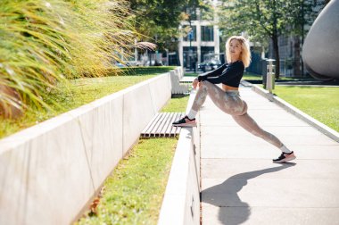 Strong sportswoman in training clothes warming up and doing stretch exercises at the street in summer day morning. Sport concept 