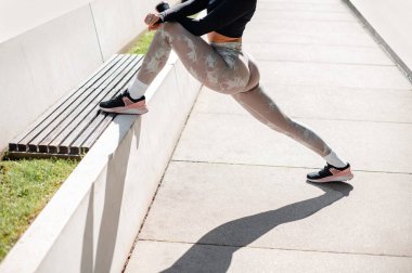 Cropped view of the strong sportswoman in training clothes warming up and doing stretch exercises at the street in summer day morning. Sport concept 