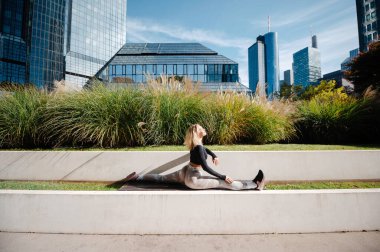 Blonde woman doing leg-split and warming up muscles at the sunny street alone. Sport and recreation concept 