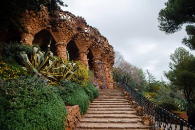 Barcelona 'daki Gaudi parkındaki güzel sütunlara giden merdiven. İspanya 'da egzotik yeşil çalıların ve çiçek bitkilerinin yanında taş kemerler..