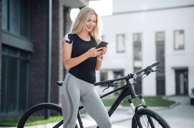 Aktif kıyafetli gülümseyen genç bir kadın elinde modern akıllı telefonuyla siyah bir bisikletin yanında duruyor. Kafkasyalı sarışın internette sörf yapıyor. Sabahtan akşama kadar dinleniyor. Açık havada bisiklet sürüyor..