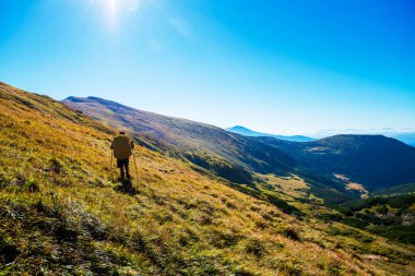 Güneşli bir havada yeşil dağlarda büyük sırt çantalı bir erkek yürüyüşçünün arka görüntüsü. Etkin zaman harcamaları sırasında büyüleyici görünüm.