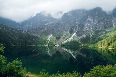Koyu bulutlar ve gölde yansıması olan yeşil ormanla kaplı güzel sisli dağlar. Taşlı sahil. Morskie Oko. Denizci gözü. Yüksek Tatras, Zakopane, Polonya