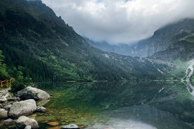 Koyu bulutlar ve gölde yansıması olan yeşil ormanla kaplı güzel sisli dağlar. Taşlı sahil. Morskie Oko. Denizci gözü. Yüksek Tatras, Zakopane, Polonya