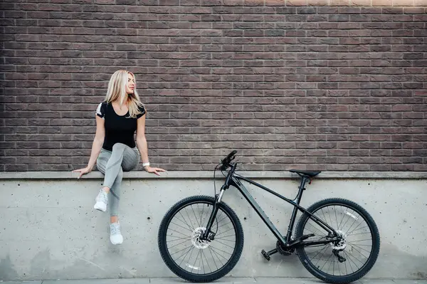 朝のワークアウトの後に休んでいる間 黒い自転車の近くの通りに座って金髪の髪を持つ美しい白人女性 屋外活動後のリラクゼーション サイクリングのコンセプト — ストック写真
