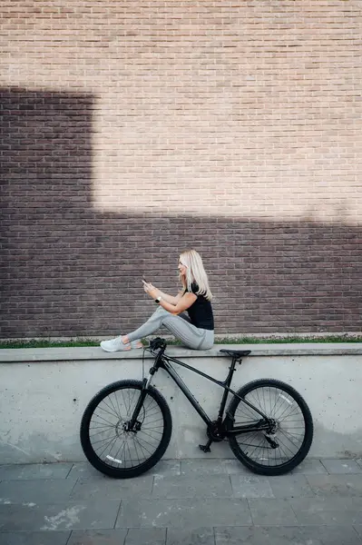 黒い自転車の近くに屋外に座って 現代のスマートフォンを使用して運動体形状を持つ魅力的な女性 スポーツウェアの白人女性は 新鮮な空気でトレーニングした後休憩を取ります — ストック写真