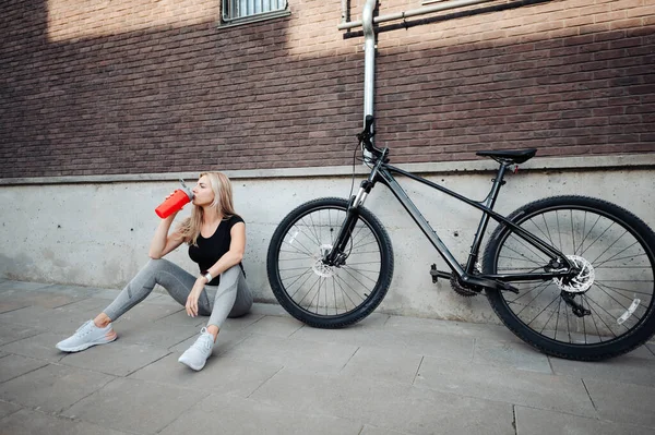 アウトドアで座ってスポーツボトルから水を飲むアクティブウェアで魅力的な白人女性 朝のサイクリングの後に若いブロンドがさわやかな 近くに立つ黒い自転車 — ストック写真