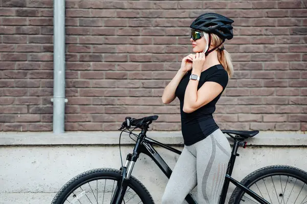黒い自転車の近くに立ち 眼鏡をかけてヘルメットをかぶっている白人女性の側面図 新鮮な空気をサイクリングしながら安全装置を使用してスポーティブロンド — ストック写真