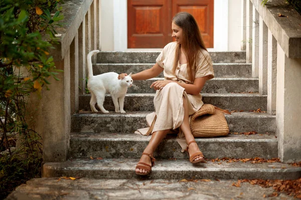 通りの階段に座り 足を横切って白いホームレスの猫を飼っている若い快適な女性 かわいい動物と遊ぶ軽い服装と茶色のサンダルで服を着た優しい笑顔の女性 — ストック写真